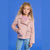 A young girl with curly blonde hair stands against a solid blue studio background wearing a light pink faux-leather moto jacket over a purple patterned top and light blue jeans.