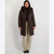 A woman modeling a long dark brown puffer coat with a large hood, worn over a dark top and paired with beige wide-leg trousers and black shoes, against a plain light gray studio background.