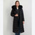 A woman modeling a long black puffer coat with a large black fur-trimmed hood, standing against a plain light gray studio background.