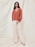 Woman wearing a rust-colored blouse and beige pants against a white curtain backdrop