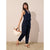 A woman models a navy blue sleeveless wrap-style jumpsuit with cropped pants, holding a cream and brown handbag against a textured beige wall and sandy floor.