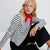 A young girl with blonde hair sits against a white background, wearing a black and white horizontally striped long-sleeve top, black wide-leg pants, and a red neck scarf.