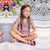 A young girl with long light brown hair sits cross-legged on white steps, wearing a pink leopard-print short-sleeve top, pink sandals, and pink-framed glasses. A mirrored floral arrangement is visible on the left side of the background.