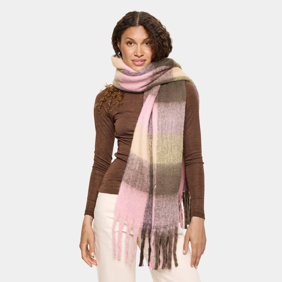 A woman modeling a thick, soft striped knit scarf in shades of pink, beige, brown, and gray, paired with a brown long-sleeve top and white pants against a light gray studio background.