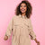 A woman with curly brown hair smiling and wearing a beige utility shirt dress with a drawstring waist, set against a solid pink background.