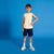 A young boy stands against a solid blue studio background wearing a sleeveless tank top with yellow and white horizontal stripes, navy blue shorts, white socks, and light-colored sneakers.