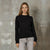 A woman modeling a black long-sleeve crewneck sweater with small silver rhinestone embellishments across the chest, paired with light gray jeans, set against a textured concrete wall.