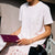 A woman wearing a white short-sleeve t-shirt with embroidered butterfly details on the chest, paired with a white lace skirt. She is holding a dark purple notebook, with a black handbag in the foreground.