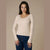 A woman modeling a ribbed long-sleeve scoop-neck sweater in cream, paired with dark blue jeans, against a solid beige studio background.