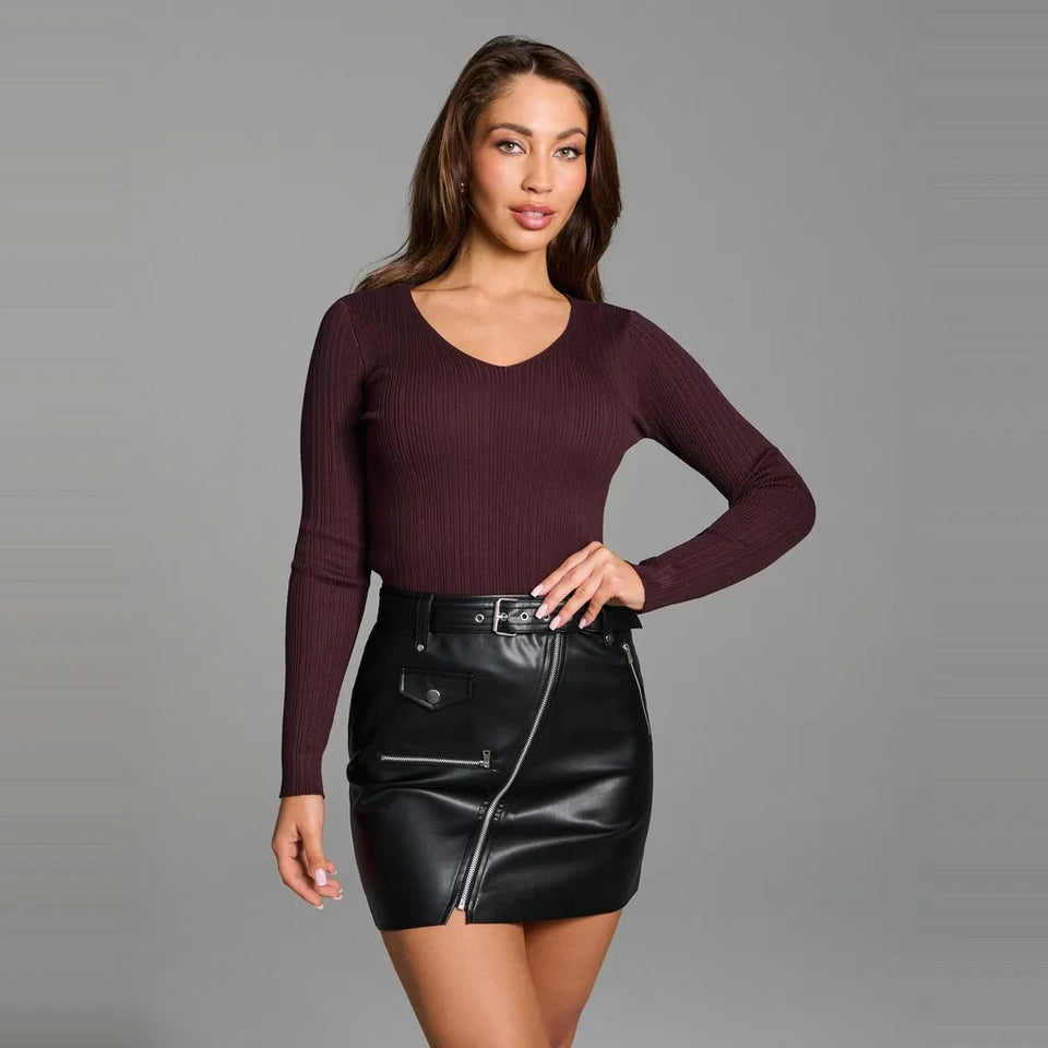A woman modeling a dark burgundy ribbed long-sleeve top and a black faux-leather mini skirt with a front zipper, photographed against a neutral gray studio background.