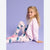 A young girl with curly blonde hair sits on a lavender studio background wearing a light pink sweatshirt and matching pink leggings with a blue cartoon character print.