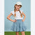 A young girl with blonde braids is wearing a white short-sleeve top with lace ruffle sleeves, a light blue tiered skirt with white floral prints, and a white baseball cap. She is posing outdoors against a light blue sky and green grass background.