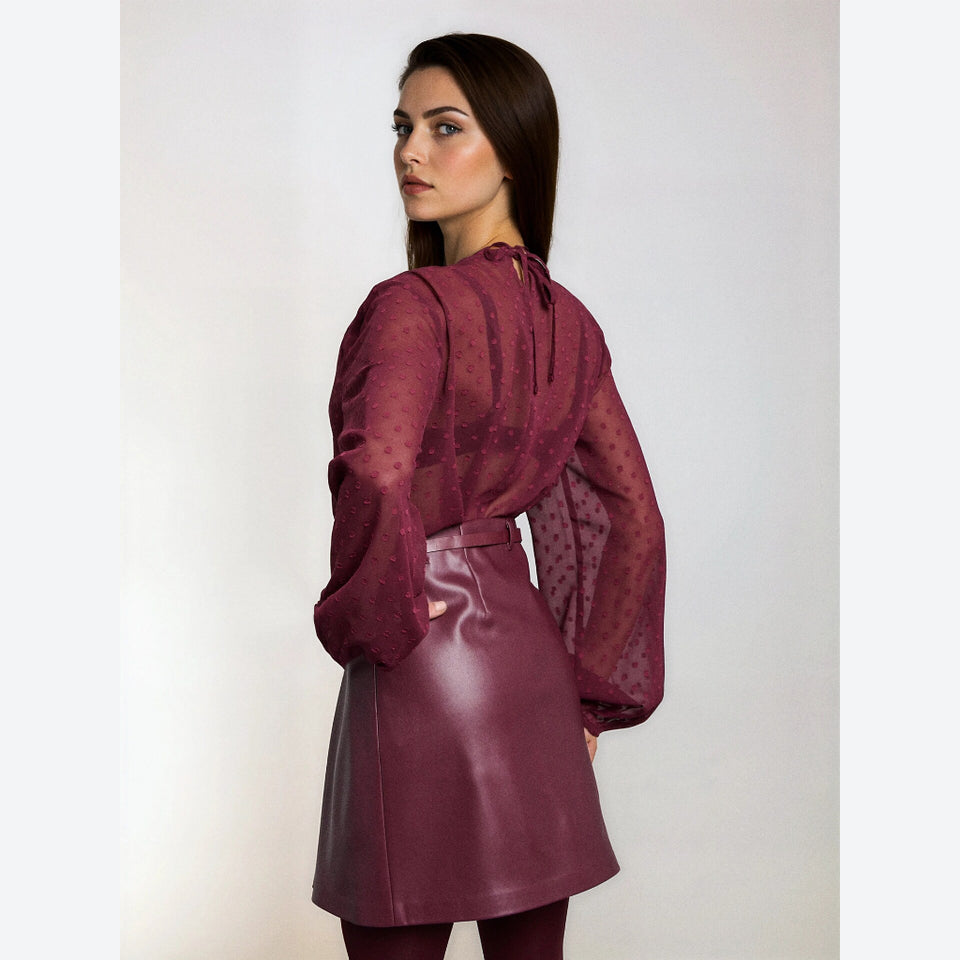 A woman modeling a burgundy sheer long-sleeve blouse with a tie-neck detail and a matching faux-leather mini skirt, shown from a three-quarters back view against a light gray studio background.