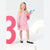 A young girl with blonde hair stands in a studio wearing a pink long-sleeve dress with small black polka dots, white socks, and black Mary Jane shoes. She is holding a large pink pencil-shaped plush toy.