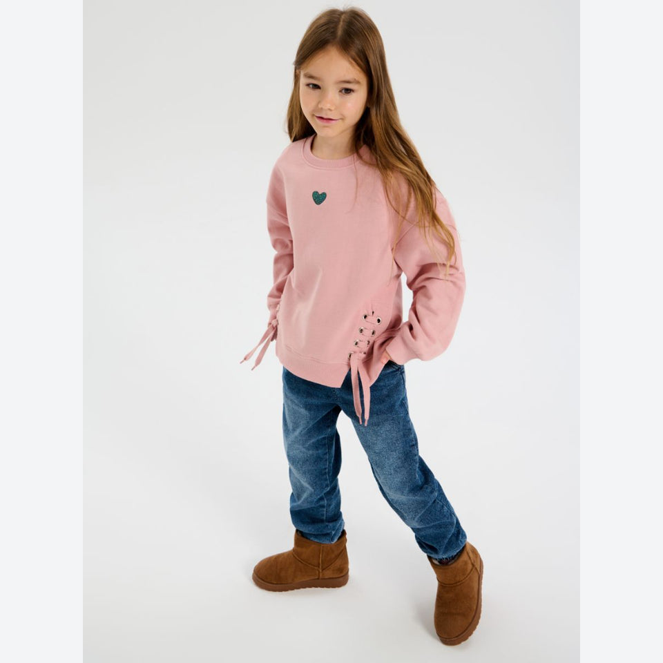 A young girl with long light brown hair stands in a studio wearing a dusty pink crewneck sweatshirt with a small green heart on the chest and lace-up tie details on the sleeves, paired with blue jeans and brown suede boots.