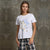 A woman modeling a white short-sleeve graphic T-shirt with a blue floral design and gold metallic 'MAGIC' lettering, paired with plaid pants, set against a textured gray concrete wall.