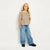 A young girl with blonde hair stands against a plain white studio background wearing a beige and brown patterned knit long-sleeve top, wide-leg blue jeans, and black shoes.