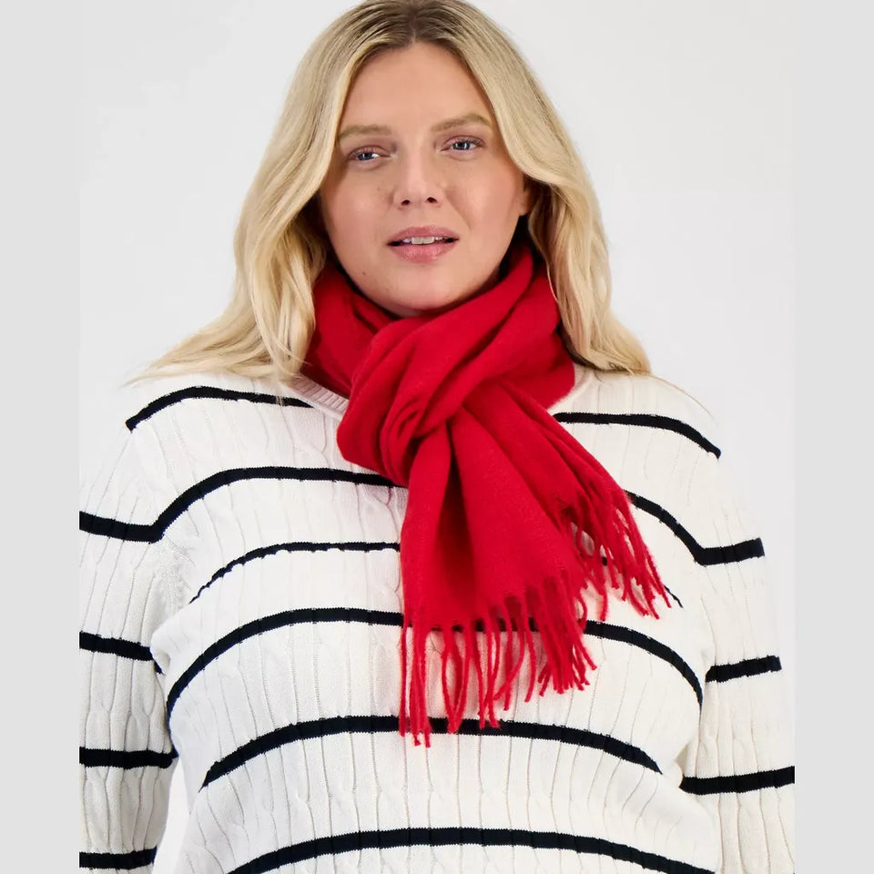 A blonde woman wearing a bright red knit scarf with fringe and a white ribbed sweater with black horizontal stripes, photographed against a light gray studio background.