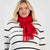 A blonde woman wearing a bright red knit scarf with fringe and a white ribbed sweater with black horizontal stripes, photographed against a light gray studio background.