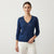 A woman modeling a navy blue long-sleeve V-neck knit sweater with a subtle textured pattern, paired with white pants, against a plain light gray studio background.