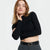 A woman wearing a black ribbed long-sleeve crop top and dark gray trousers with a black belt, posing against a light gray studio background.