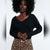A woman with a large black afro wearing a black long-sleeve V-neck top and leopard print pants, standing against a white studio background.