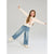 A young girl with long light brown hair is posing in a studio setting, wearing a cream-colored long-sleeve hoodie with a teddy bear graphic, wide-leg blue jeans, and cream sneakers. She is captured mid-motion with arms outstretched against a solid light gray background.