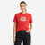 A woman modeling a red short-sleeve crewneck t-shirt with a retro Nike graphic on the chest, paired with black jeans, against a plain white background.