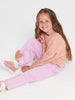 Young girl wearing a pink sweatshirt and light purple pants sitting on a white background