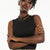 A woman with short curly black hair wearing a black sleeveless mock-neck top and a leopard print skirt, accessorized with gold hoop earrings and layered gold necklaces, posing with crossed arms against a plain white background.