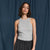 A woman modeling a light gray ribbed sleeveless tank top paired with dark gray pinstriped trousers, standing against a dark blue draped fabric backdrop.