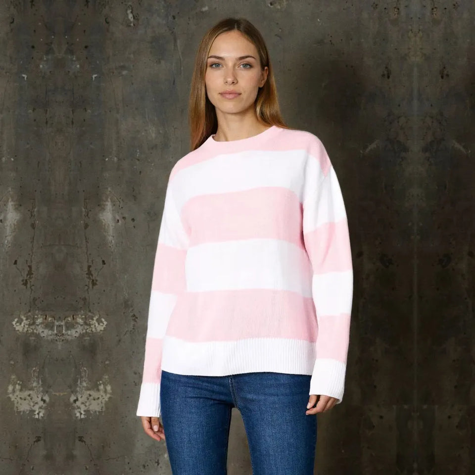 A woman wearing a pink and white horizontally striped knit sweater with blue jeans, standing against a dark textured concrete wall.