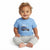 A smiling toddler sitting cross-legged on a white background, wearing a light blue short-sleeve t-shirt with a 'POLO surf club' graphic and light gray sweatpants.