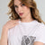 A close-up of a woman wearing a white short-sleeve crewneck t-shirt with a black and white snakeskin print graphic on the front pocket area, set against a plain light gray background.