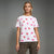 A woman modeling a white short-sleeve crewneck t-shirt with an allover pink and red lipstick kiss print, paired with leopard-print pants, against a neutral gray studio background.
