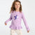 A young girl with curly brown hair wearing a lavender long-sleeve top with a purple and white sequined star on the chest, paired with cream patterned leggings, against a plain white background.