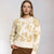 A woman modeling a cream-colored knit sweater with a textured gold floral pattern, paired with brown trousers, against a neutral light gray background.