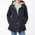 A woman wearing a dark navy hooded jacket with a beige sherpa lining, gold-tone snap and zipper details, and front patch pockets, paired with a gray shirt and blue jeans against a plain white background.