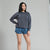 A woman modeling a dark gray washed crewneck sweatshirt with tonal embroidered text, paired with blue denim shorts, against a plain light gray studio background.