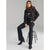 A model poses in a black tailored outfit consisting of a blazer, trousers, and a dark brown knit scarf, seated on a black metal stool against a neutral gray studio background.