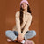 A young girl sitting cross-legged against a solid brown background, wearing a beige half-zip hoodie, light blue jeans, a pink beanie, and pink sneakers.