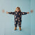 A young child stands with arms outstretched wearing a dark navy one-piece snowsuit covered in a light pink teddy bear print, paired with tan boots, against a solid light blue background.