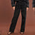 A model wearing black wide-leg jeans with a relaxed straight-to-slightly-flared leg, styled with a black puffer jacket and black strappy sandals against a solid warm brown studio background.