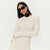 A woman wearing a textured off-white long-sleeve dress with a black fabric flower at the neckline, styled with black sunglasses and a black flower accessory, against a plain white background.