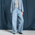 A model wearing light-wash wide-leg denim jeans, a light gray top, and a light blue denim jacket, standing against a dark blue draped fabric backdrop.