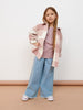 Young girl wearing a pink plaid shirt, light blue jeans, and white sneakers on a beige floor with a white wall background.