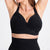 A close-up of a woman wearing a black wireless sports bra with a deep V-neckline and wide shoulder straps, paired with a matching black high-waisted shapewear or leggings bottom against a plain white background.