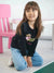 Young girl wearing a black sweatshirt with a teddy bear design, sitting on a white surface.