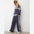 A woman modeling a navy blue tracksuit with white stripes, featuring a cropped long-sleeve top with a white chest stripe and matching wide-leg pants with a white side stripe, paired with white sneakers against a plain light gray wall.