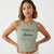 A woman modeling a ribbed sleeveless crop tank top in sage green with a dark gray 'BROOKLYN East Coast' graphic and skyline print on the front, set against a plain light gray background.